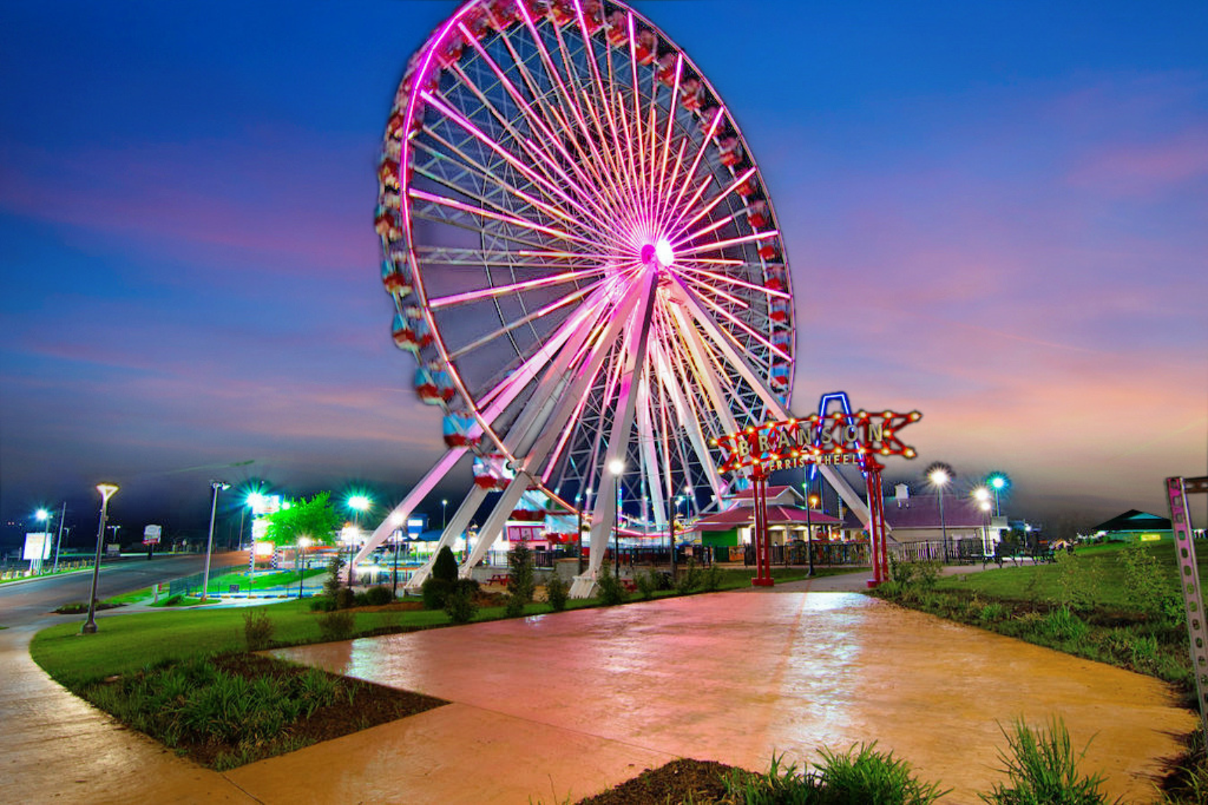 Branson Ferris wheel lit up at dusk with pink and purple lights, set against a colorful sunset sky and a paved walking path in the foreground.