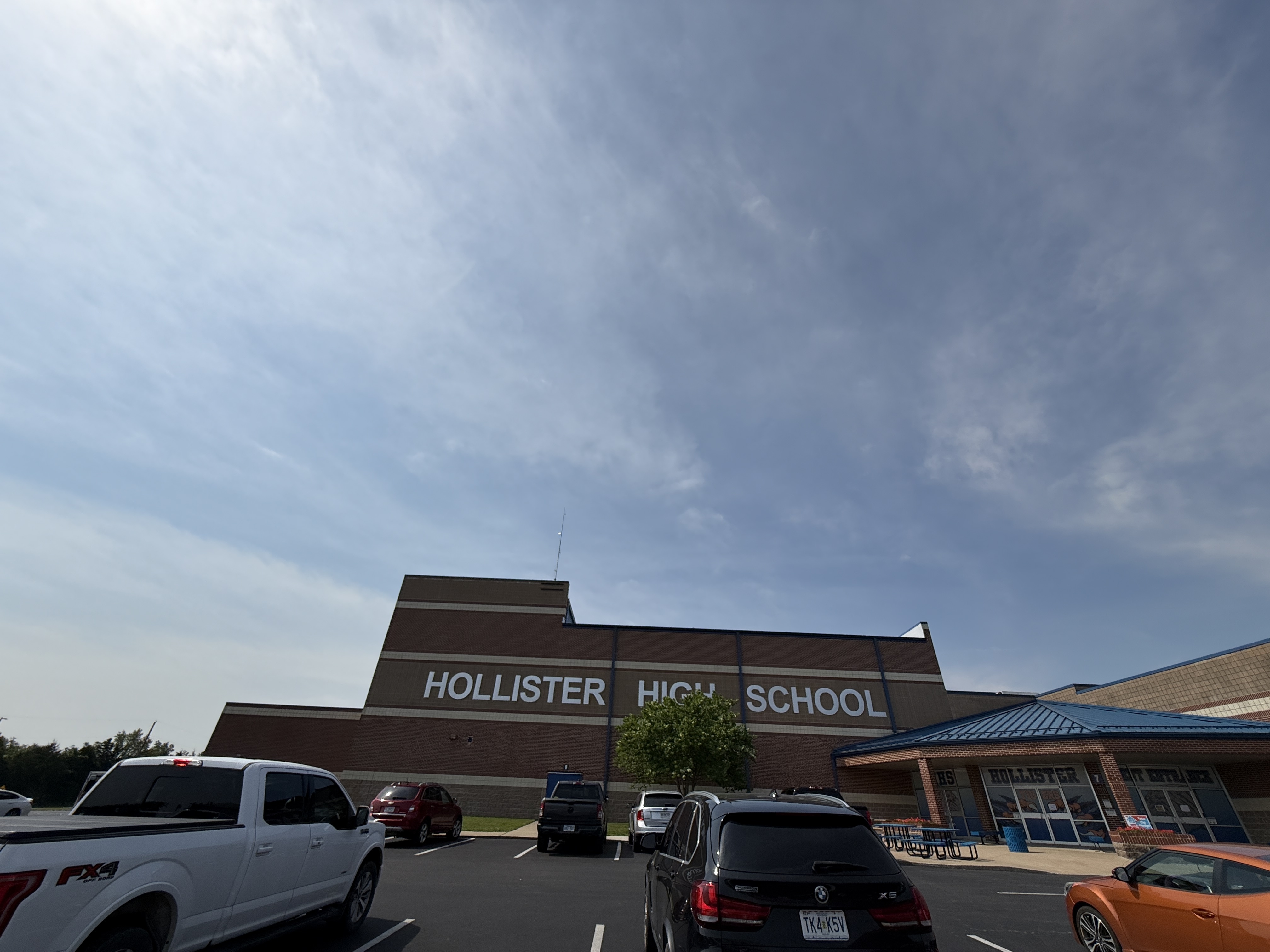 Front view of Hollister High School with parked cars in the lot and clear signage on the building, under a blue sky in Hollister, Missouri.