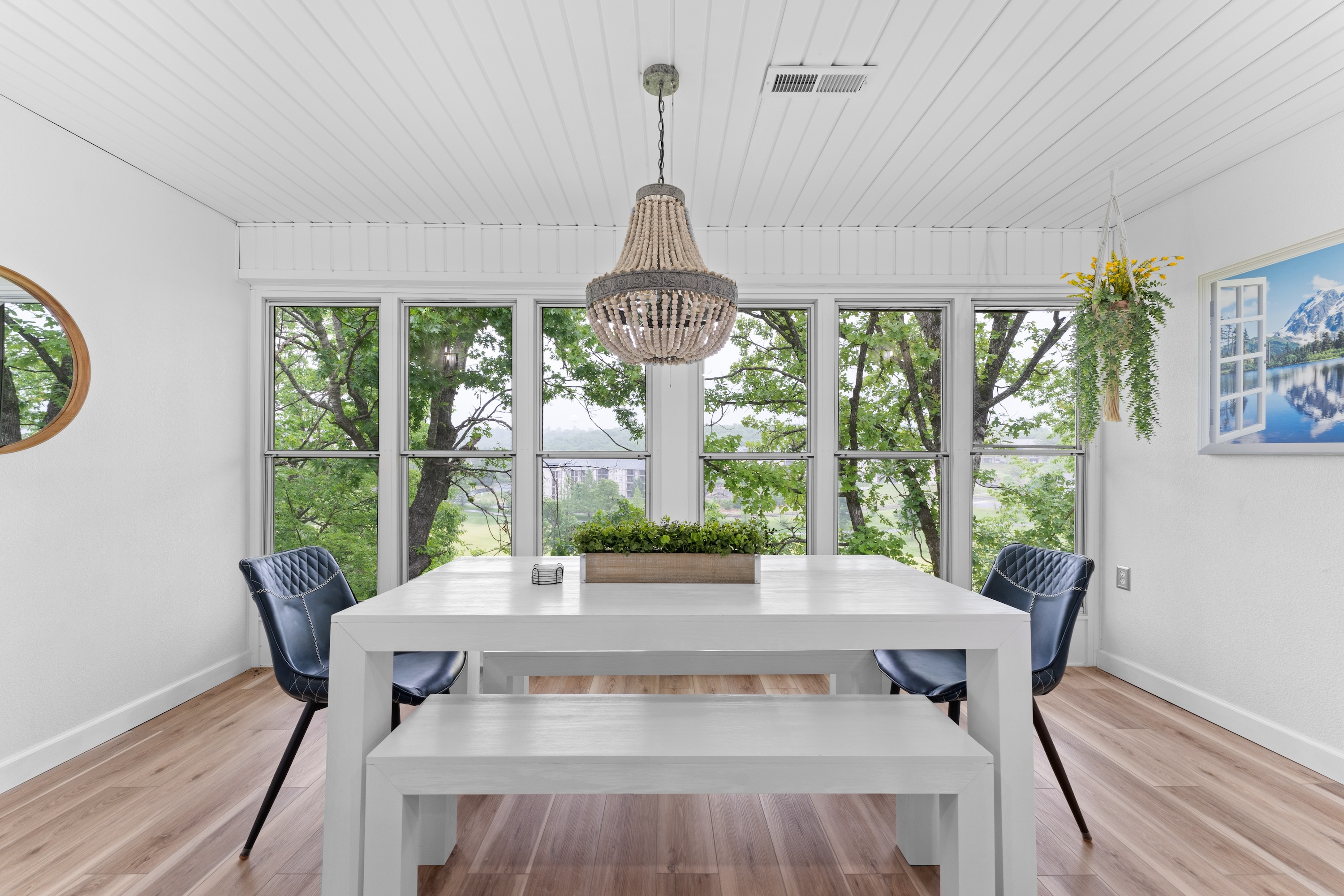 Dining area with white table, bench seating, large windows, and tree-lined views.