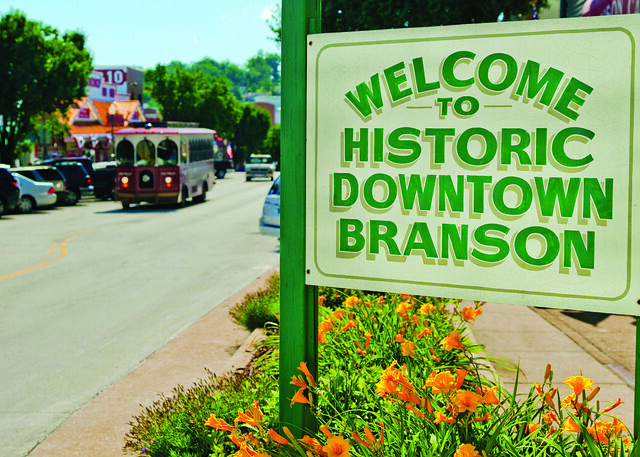 Welcome to Historic Downtown Branson sign along a street with trolley and storefronts in Branson, Missouri.