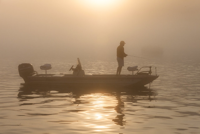 Silhouette of a person fishing from a bass boat on calm water in the early morning fog, with sunlight reflecting on the lake.