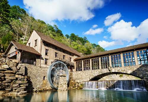 Historic watermill at Silver Dollar City in Branson Missouri with large water wheel, stone bridge, and flowing waterfall under a blue sky.
