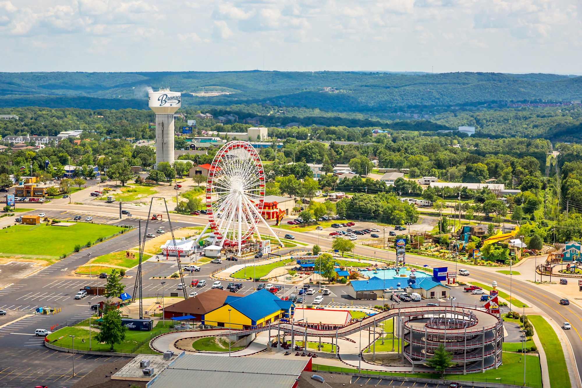 Aerial view of the Branson Strip in Missouri featuring the Branson Ferris Wheel, Highway 76, go-kart tracks, attractions, and the Branson water tower with Ozark hills in the background.