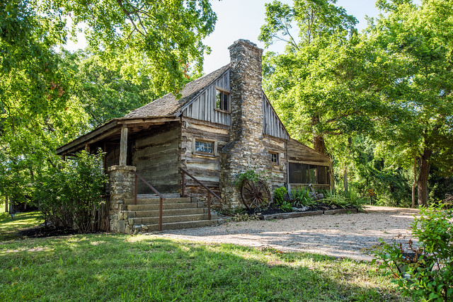 Historic log cabin with stone chimney surrounded by trees in Branson Missouri, showcasing Ozark heritage architecture and natural landscaping