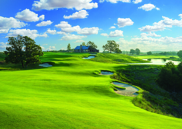 Rolling Ozark golf course with bright green fairways, sand bunkers, and water features under a blue sky with scattered clouds.