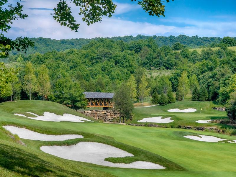 Ozark golf course with rolling green fairways, white sand bunkers, and a covered wooden bridge set against wooded hills under a blue sky.
