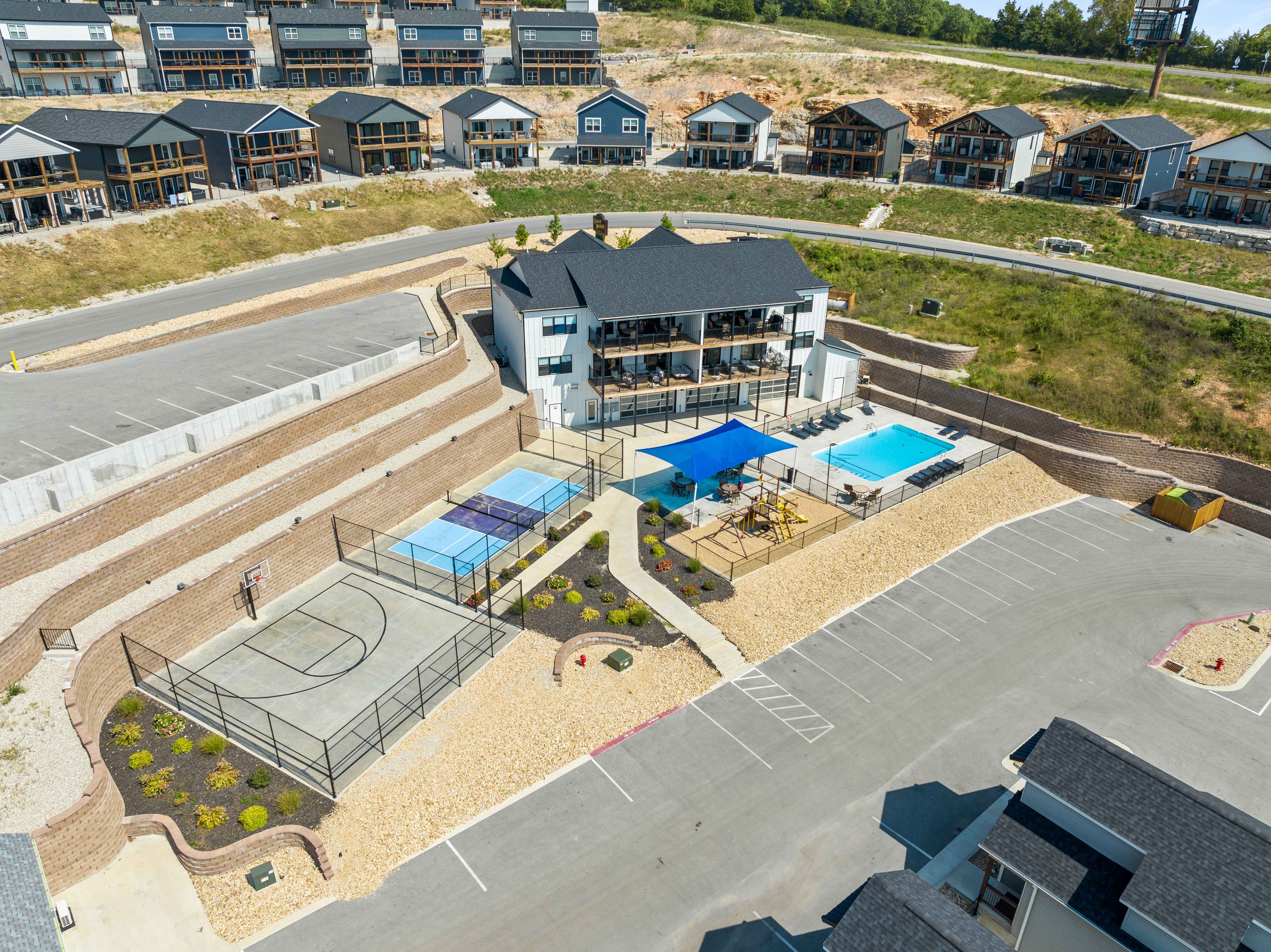 Aerial view of a residential complex with a central clubhouse building overlooking outdoor amenities. In front of the building are a fenced basketball half-court, two fenced pickleball courts, a shaded playground, and a rectangular swimming pool with lounge chairs. Curved retaining walls and landscaped paths surround the area, with a large parking lot in the foreground and rows of townhomes on the hillside behind.
