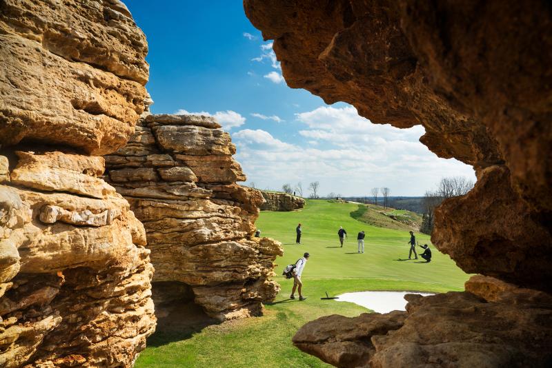 Golfers putting on a green framed by large natural rock formations in the Ozarks under a bright blue sky.