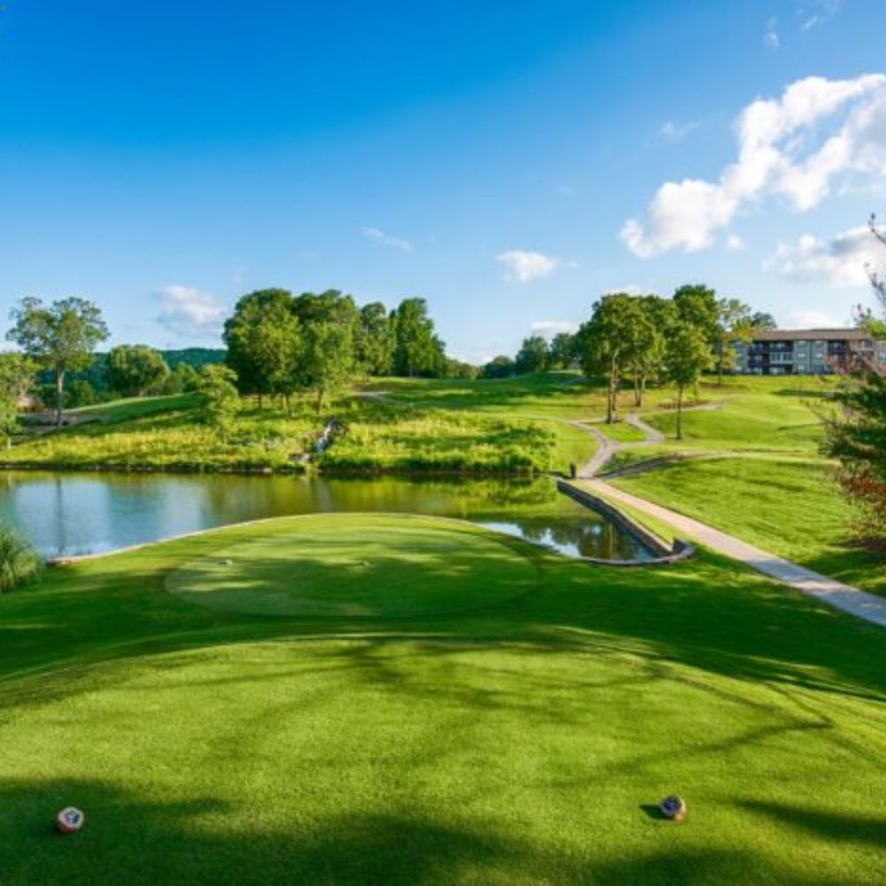 Scenic golf course in Branson, Missouri with manicured green, water hazard, walking paths, and rolling Ozark hills under a blue sky.