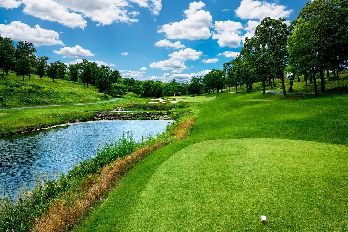 Scenic view of Thousand Hills Golf Course in Branson Missouri featuring manicured green fairway, water hazard, walking path, and rolling Ozark hills under a blue sky.