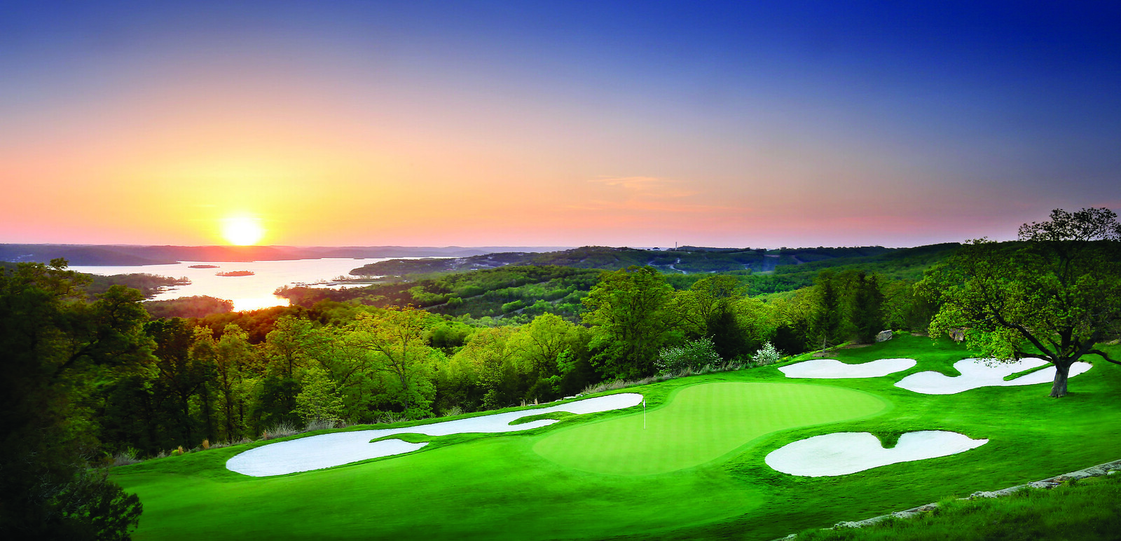 Scenic golf course in the Ozarks at sunset with manicured green, white sand bunkers, and Table Rock Lake glowing in the background under a colorful sky.